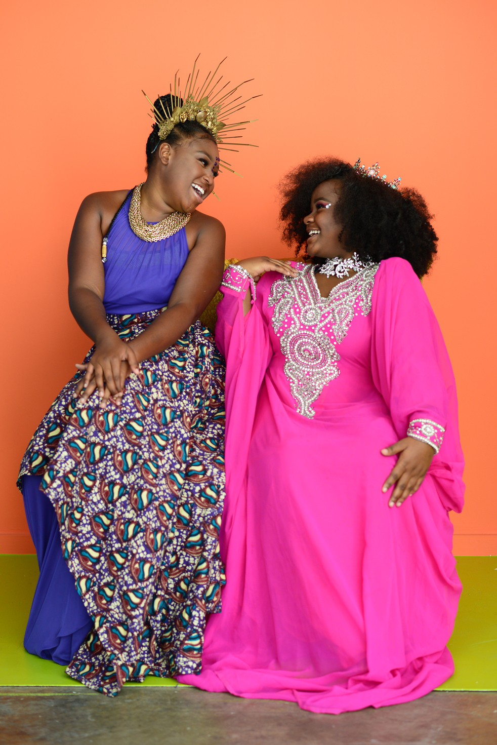 African American teen girls in royal regalia smiling at each other and sharing a throne. The girl on the left wears a royal purple dress and the girl on the right wears a hot pink gown. Both girls don crowns and sit in front of an orange backdrop. Building Opportunities & Opening Minds, 2022.