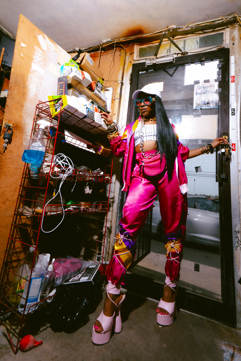 African American Woman, named Jamie LeShae Jenkins, wearing a hot pink track suit, 1990s style, posing inside of the door frame of a convenience store in Oak Cliff, Dallas, Texas. She is the founder and executive director of BOOM, a Dallas-based nonprofit. Alternate names: J LeShae, J LeShaé, Ms. J, Jamie LeShaé Jenkins.